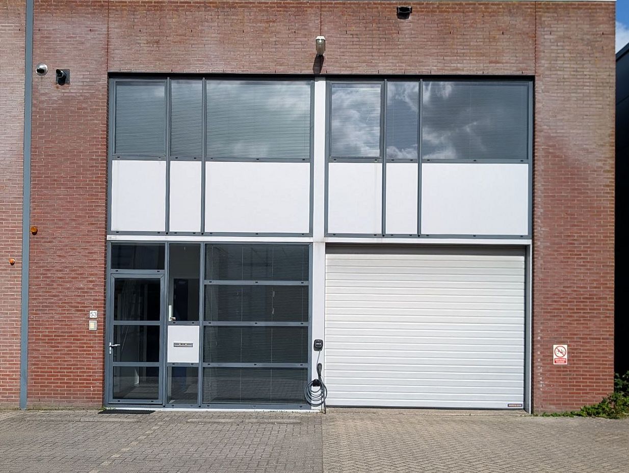Voorgevel van De Stuwdam 53: rode baksteen met een glazen entree links, een witte overheaddeur rechts en een glazen gevelstrook erboven; ervoor klinkerbestrating.