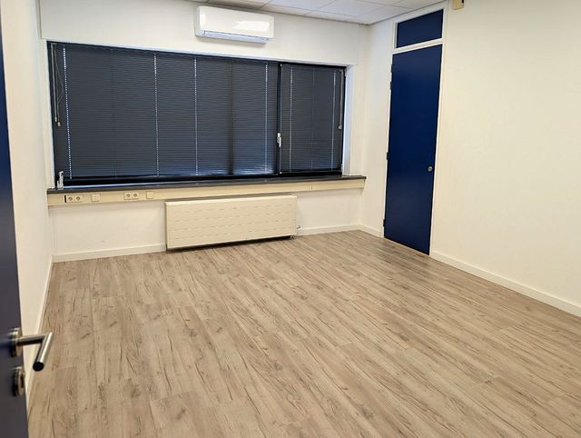 Kantoor met laminaatvloer, een brede raampartij met lamellen en een radiator, een airco-unit boven het raam en rechts een blauwe deur.
