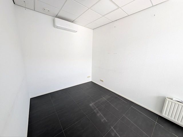 Kleine kantoorruimte met witte wanden, een donkere tegelvloer, een airco-unit aan de wand en een radiator.