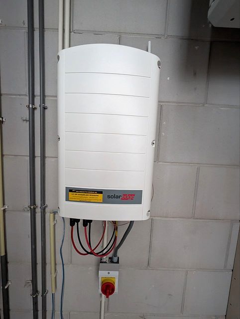 Witte omvormer aan een wand van kalkzandsteen, met kabels en een rode schakelaar eronder.