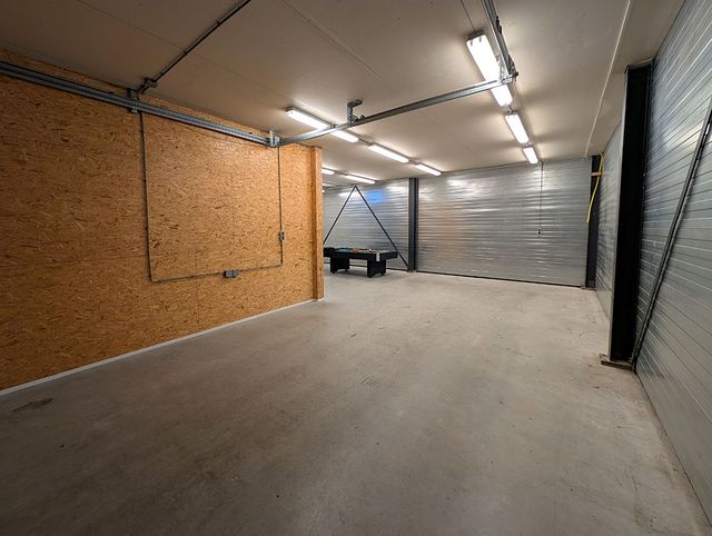 Lege opslag- en werkruimte met betonvloer en tl-verlichting, links een houten plaatwand en rechts metalen wanden.